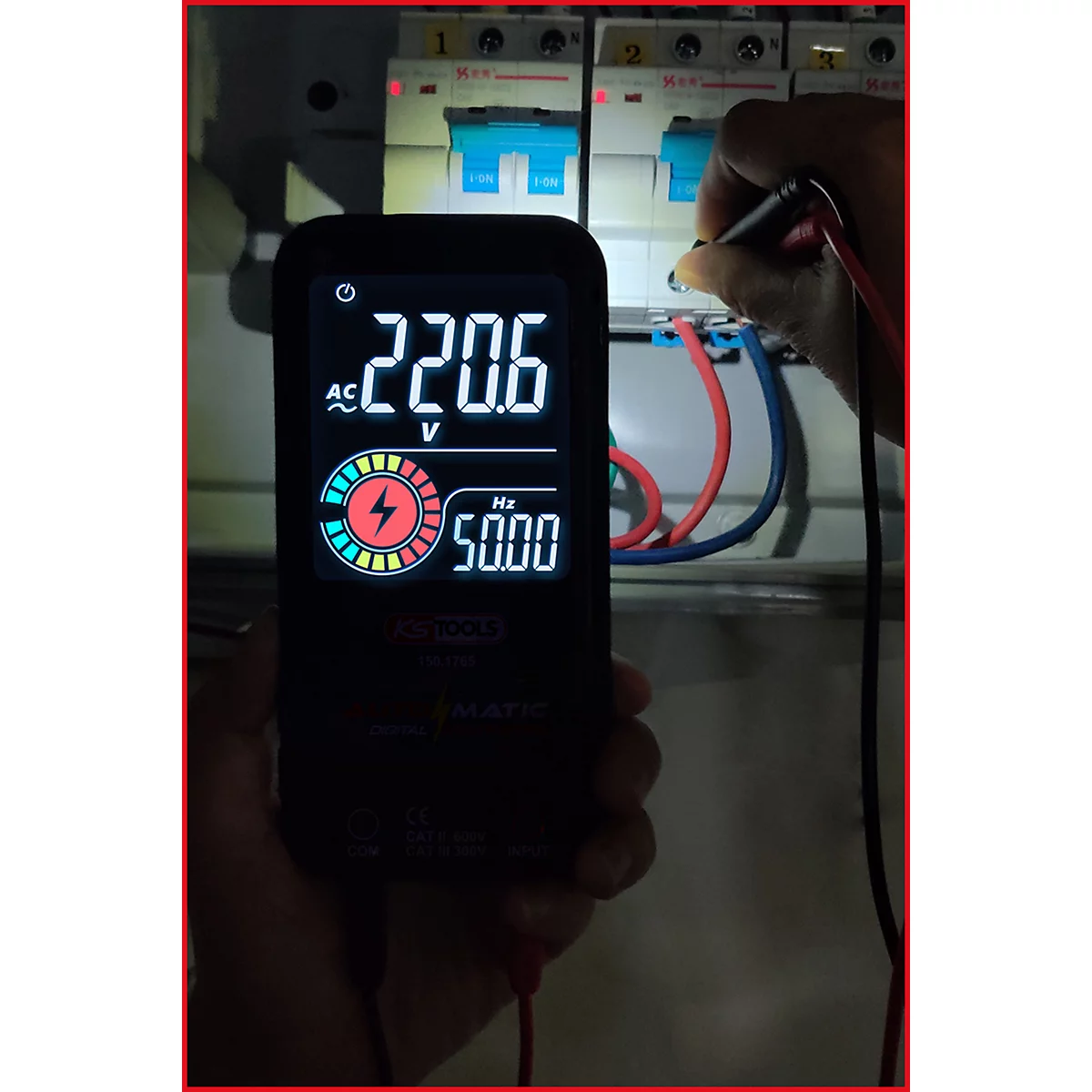 Multimeter zeigt 220,6 V und 50,00 Hz. Eine Hand berührt einen Stromkreis. Im Hintergrund sind Sicherungsautomaten zu sehen.